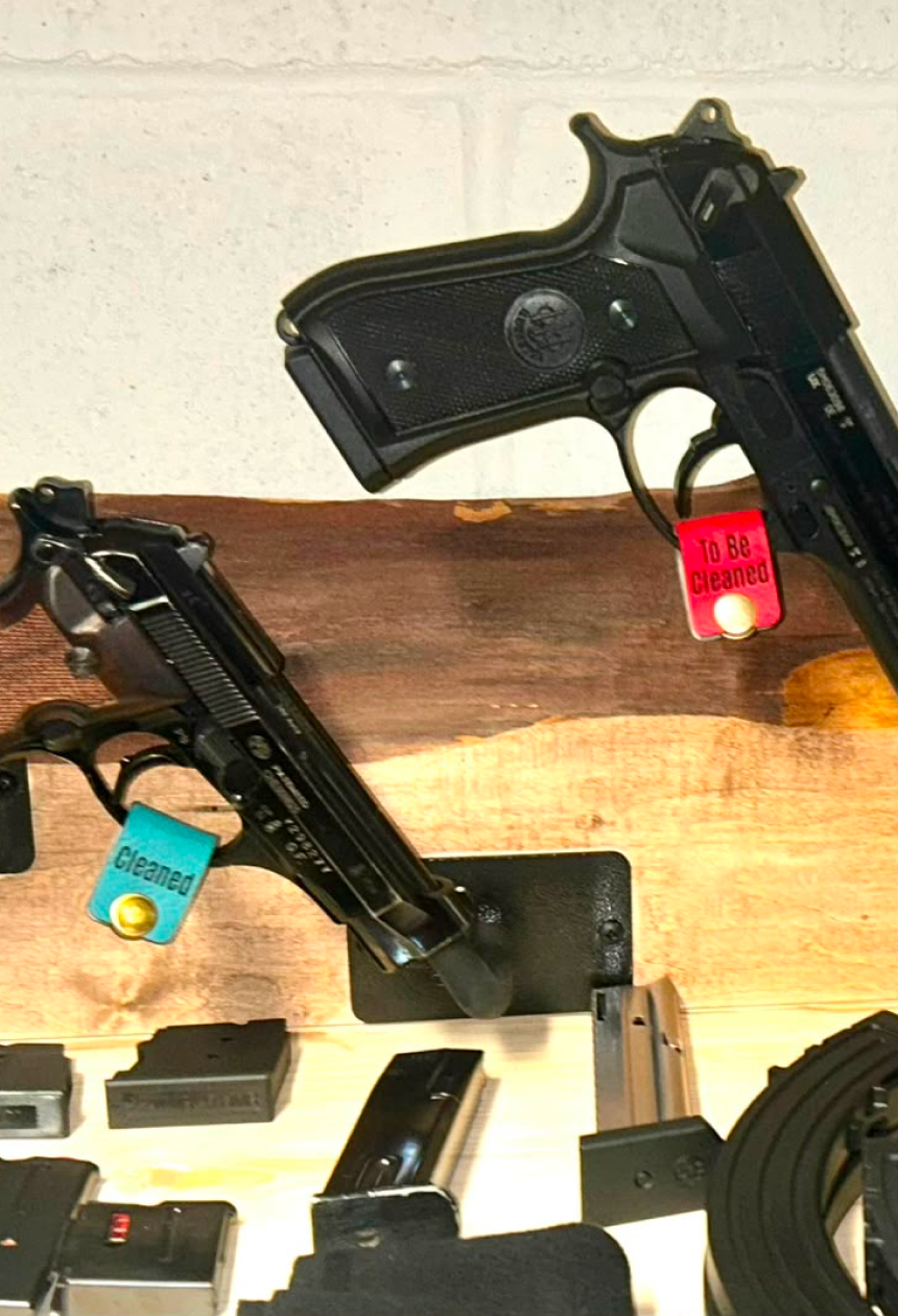 Several black handguns displayed on small stands along a wooden shelf, with red and green firearm trigger tags attached to indicate different firearm status, alongside black accessories and magazines arranged below against a light wall