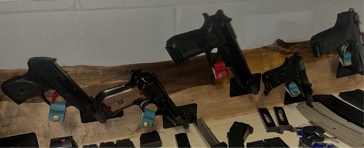 Several black handguns displayed on small stands along a wooden shelf, with red and green firearm trigger tags attached to indicate different firearm status, alongside black accessories and magazines arranged below against a light wall