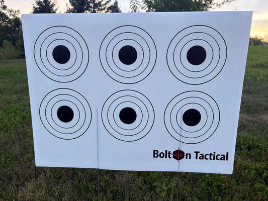 A white Bolt on Tactical shooting target sheet with six black bullseye targets, photographed outdoors on a grassy field at sunset.