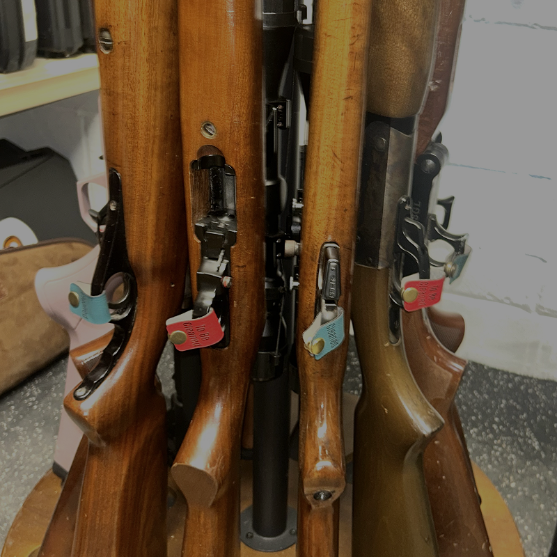 Several wooden-stocked rifles displayed vertically on a stand in a shop or storage setting, with metal lock mechanisms and small colorful tags attached.